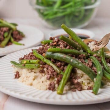 Plate with ground beef, green beans, and cauliflower rice. Fork on the plate, garlic cloves nearby, and a bowl of green beans in the background.
