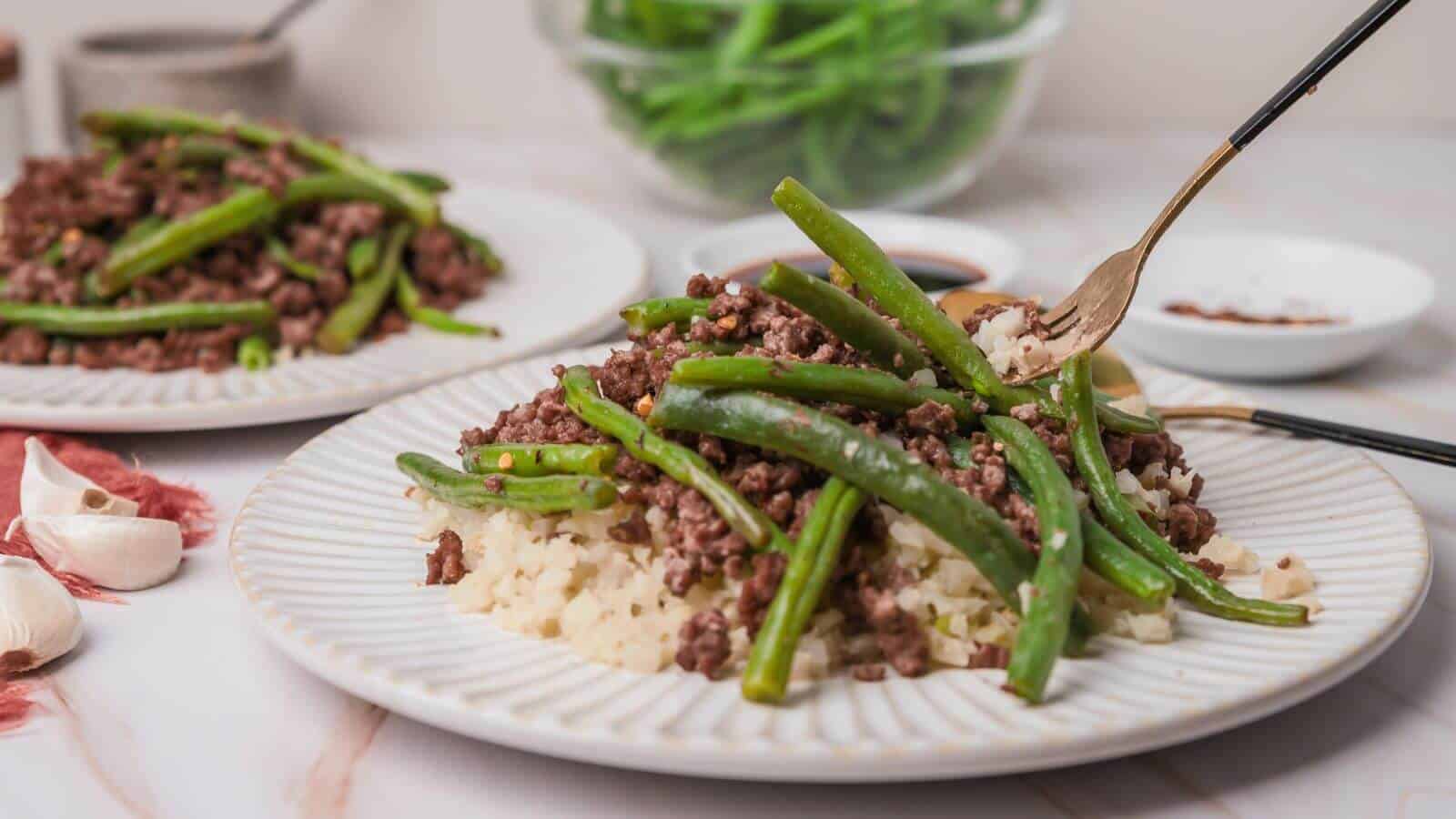 Plate with ground beef, green beans, and cauliflower rice. Fork on the plate, garlic cloves nearby, and a bowl of green beans in the background.