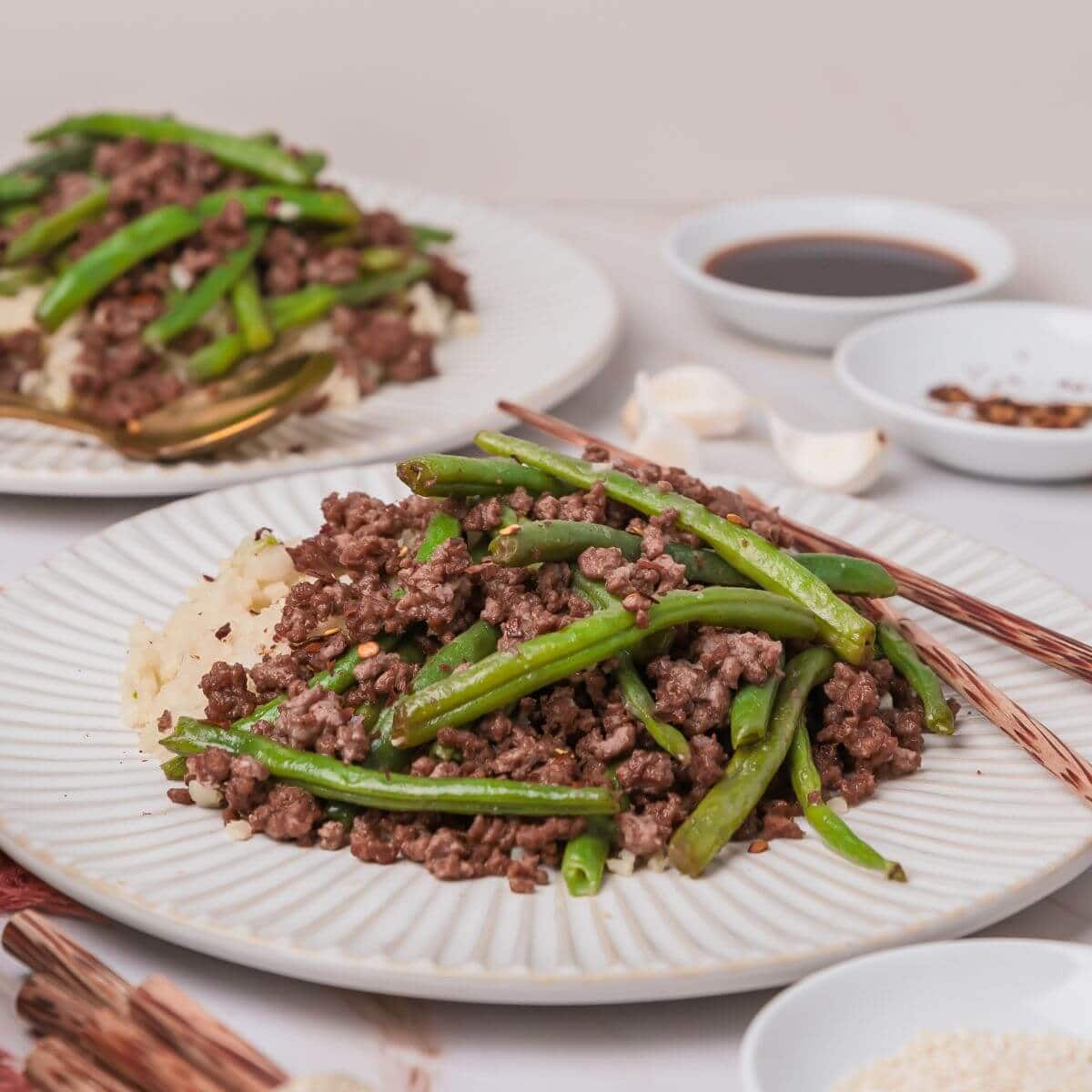 A plate of cooked minced beef with green beans on rice. Another similar dish is in the background. There are soy sauce, garlic, and spices in small bowls nearby.