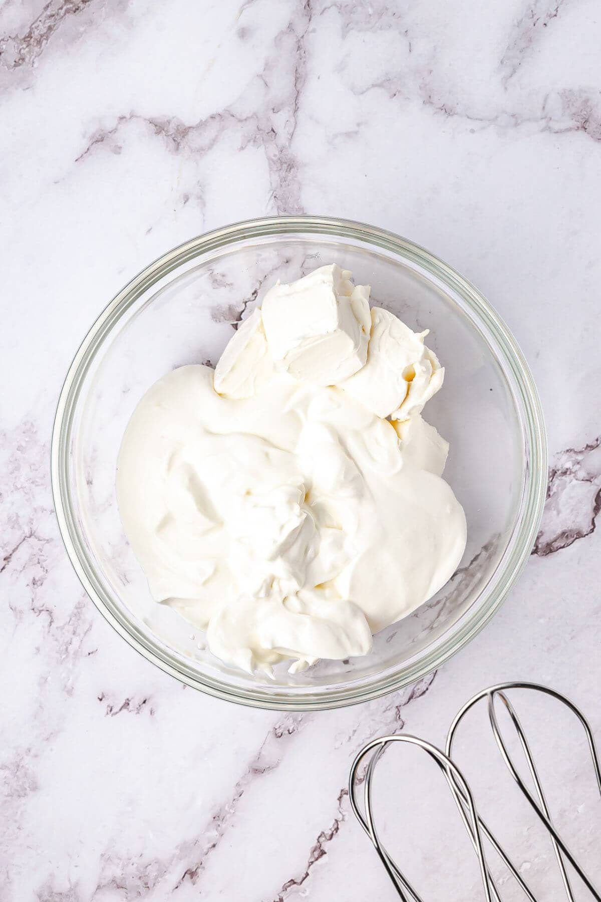 A glass bowl with a mixture of cream cheese and whipped cream on a marble surface, next to a metal whisk.