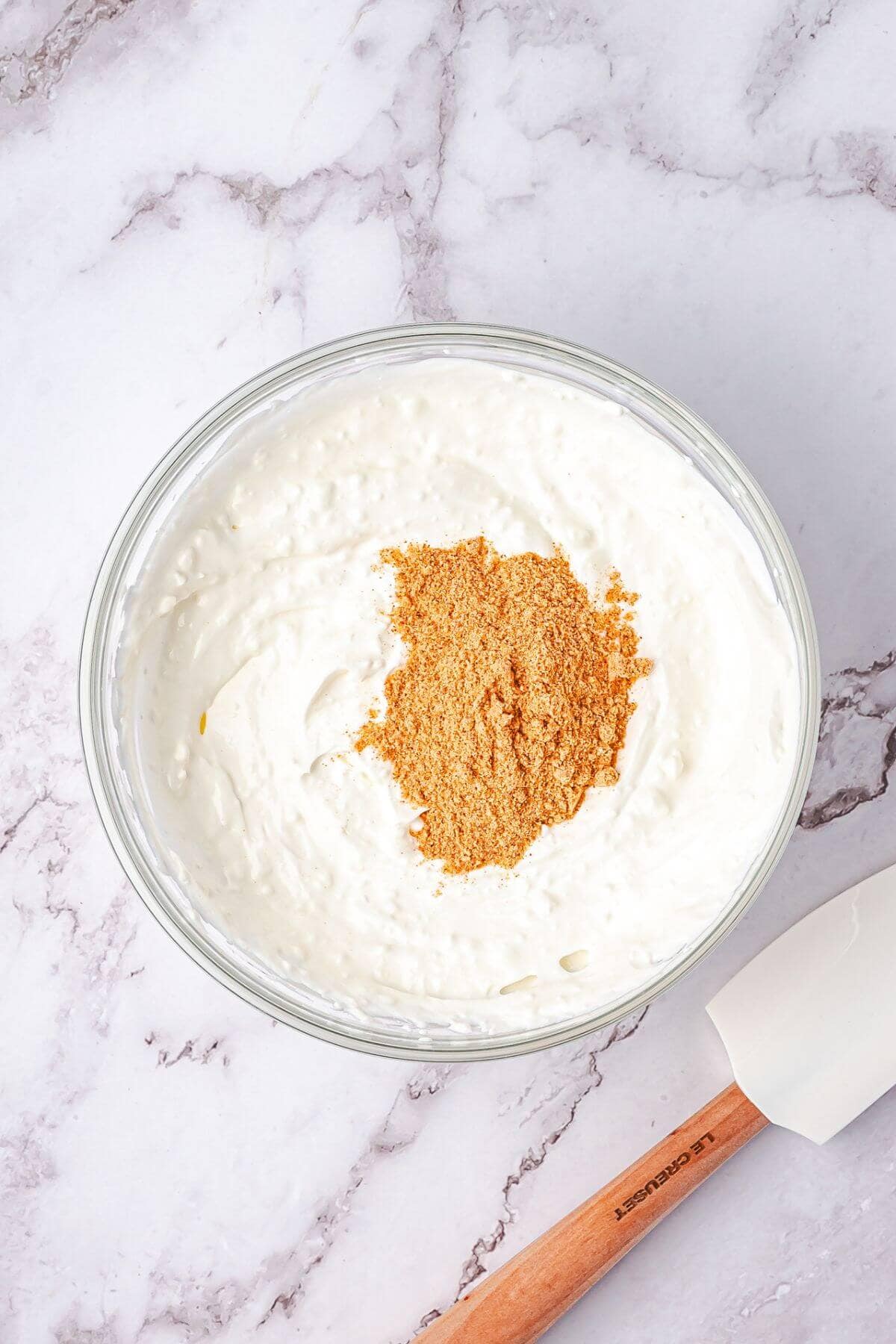 A glass bowl filled with creamy white mixture topped with a pile of brown crumbs, next to a white spatula on a marble surface.