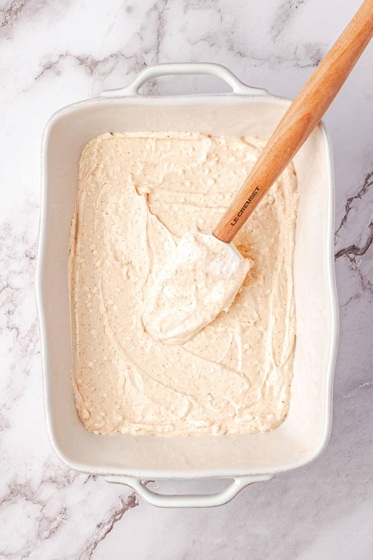 A white ceramic baking dish filled with a creamy, light tan batter being spread evenly with a wooden spatula on a marble surface.
