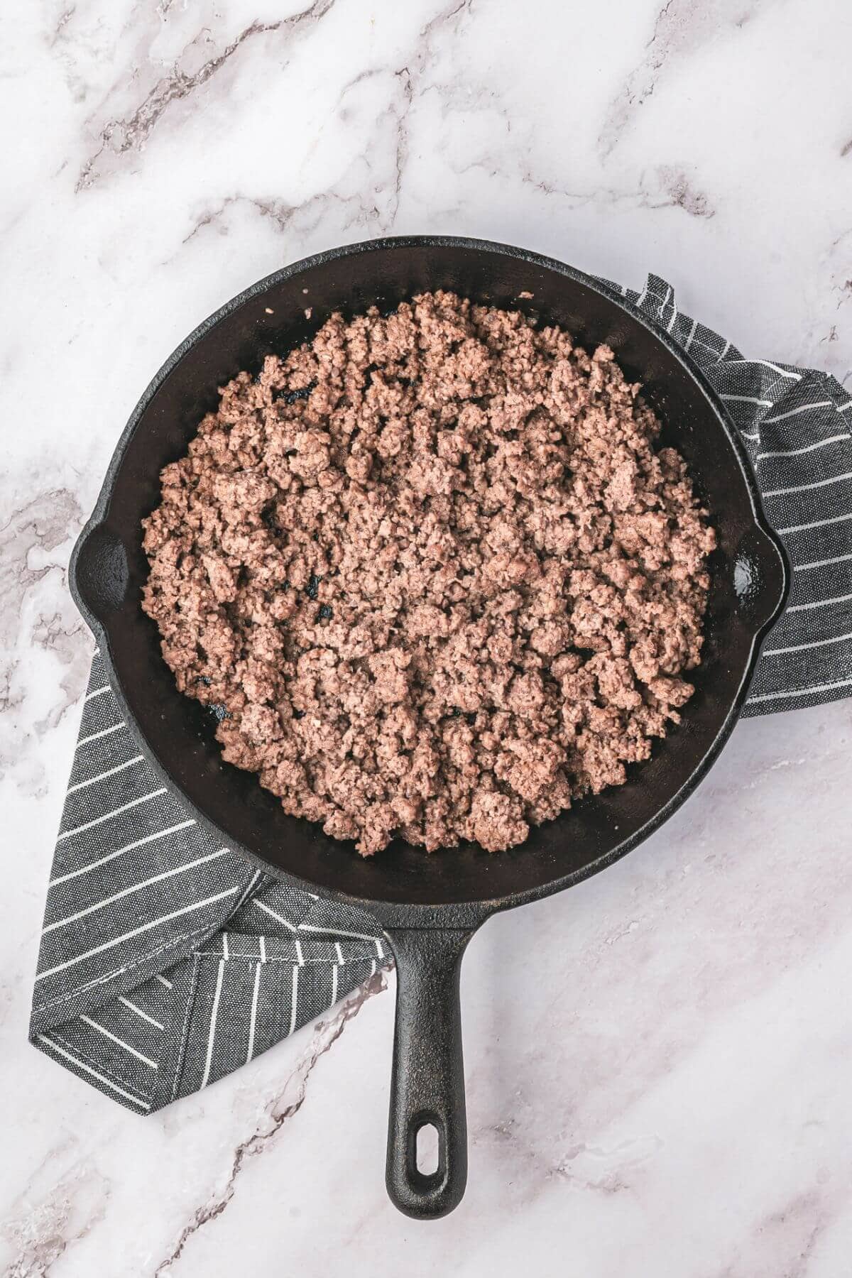 Cooked ground beef in a cast iron skillet, resting on a gray striped kitchen towel atop a light marble surface.