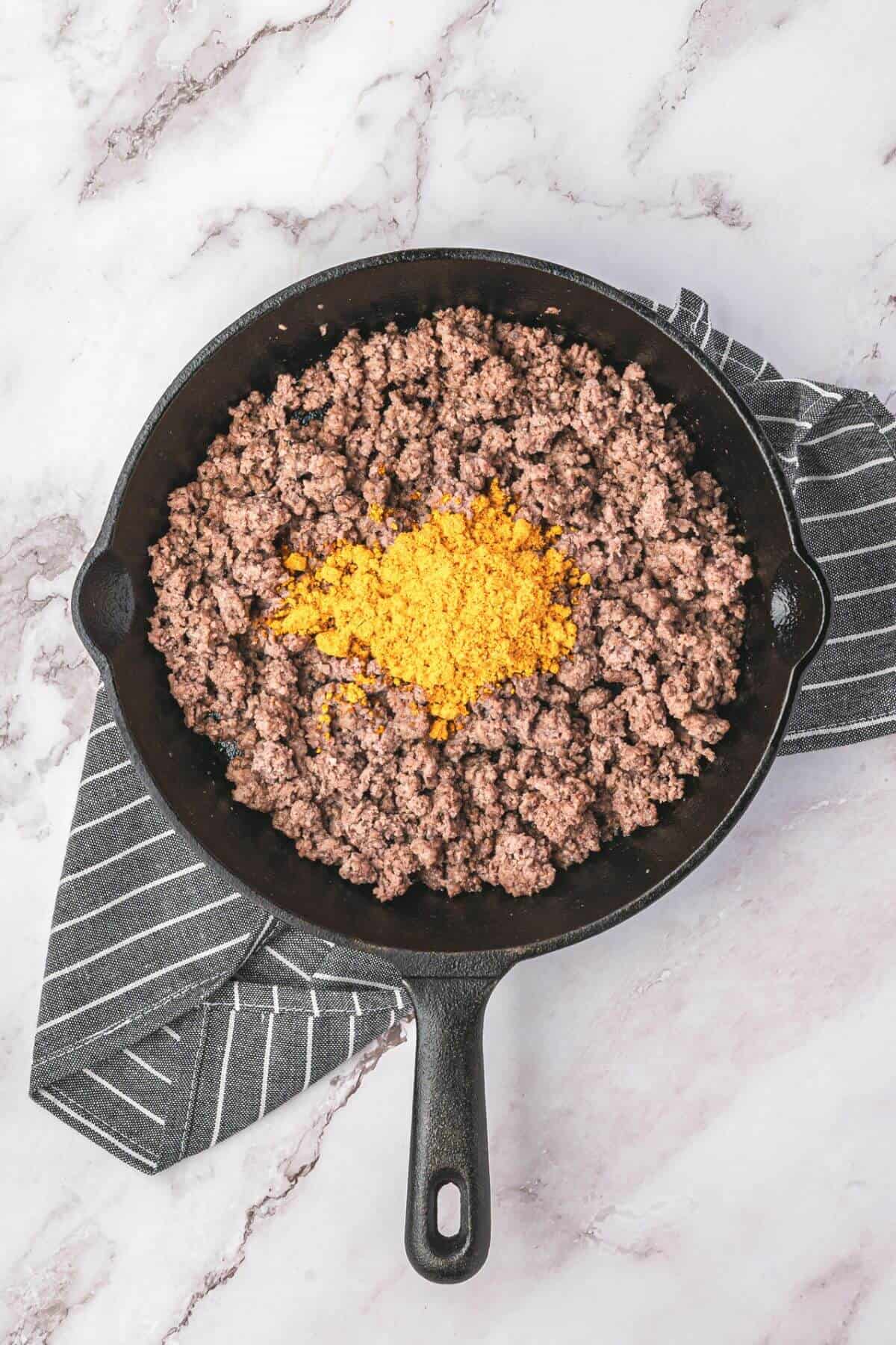A skillet filled with cooked ground beef topped with a mound of taco seasoning, placed on a striped kitchen towel on a marble surface.