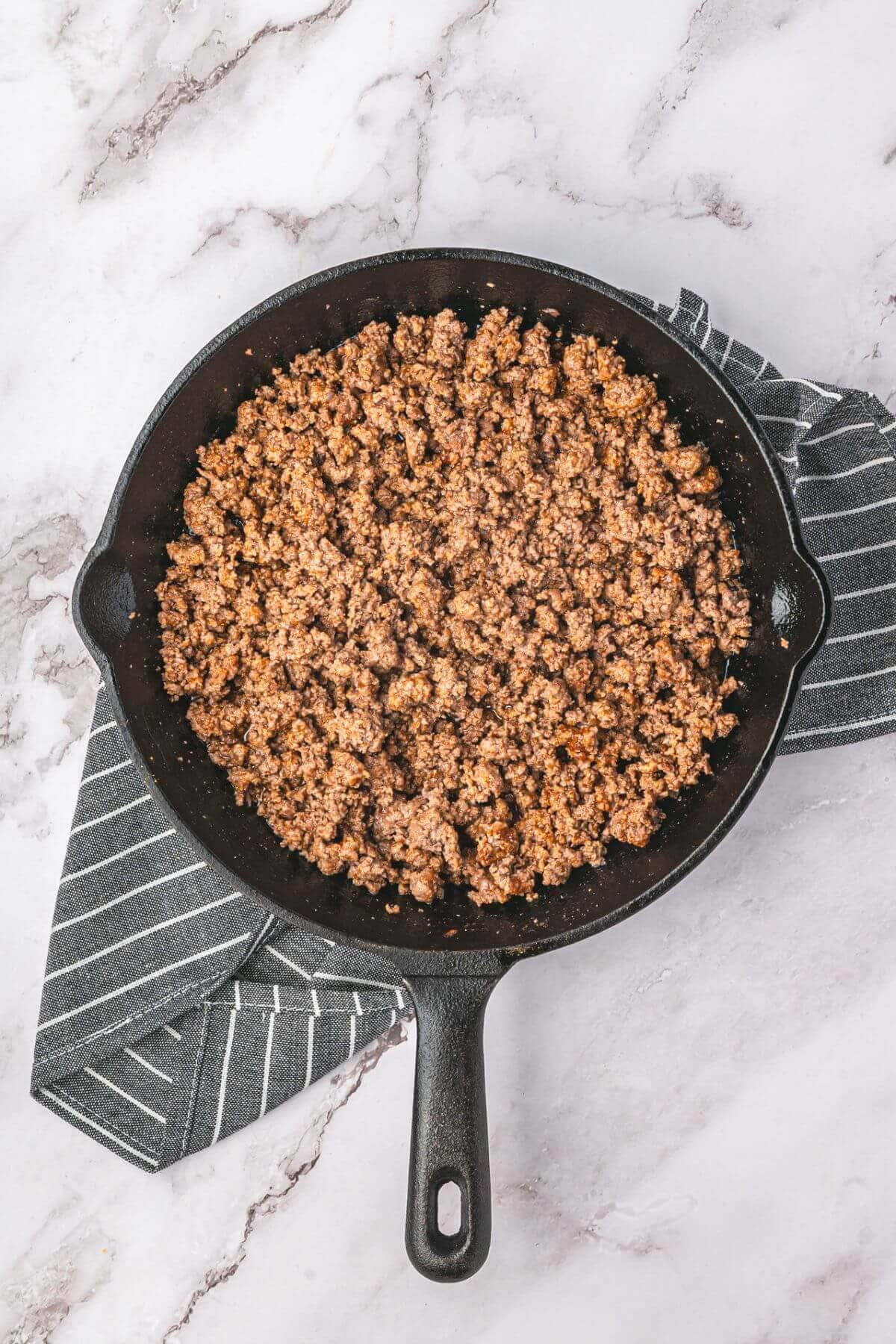 Cooked ground beef in a cast iron skillet, placed on a striped kitchen towel on a marble countertop.