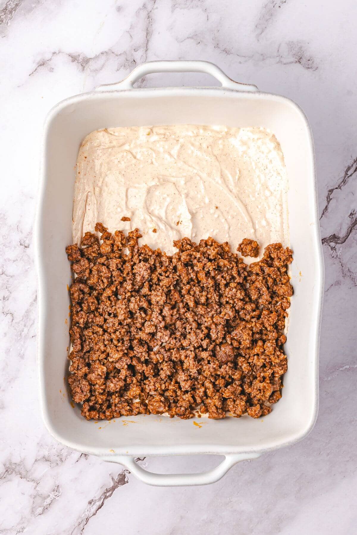 Rectangular baking dish with a layer of creamy sauce topped halfway with cooked ground meat on a marble surface.
