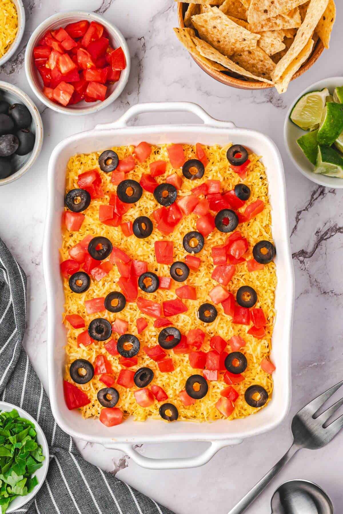 A casserole dish filled with cheesy dip topped with sliced black olives and diced tomatoes, surrounded by bowls of chips, tomatoes, olives, limes, and herbs on a marble surface.