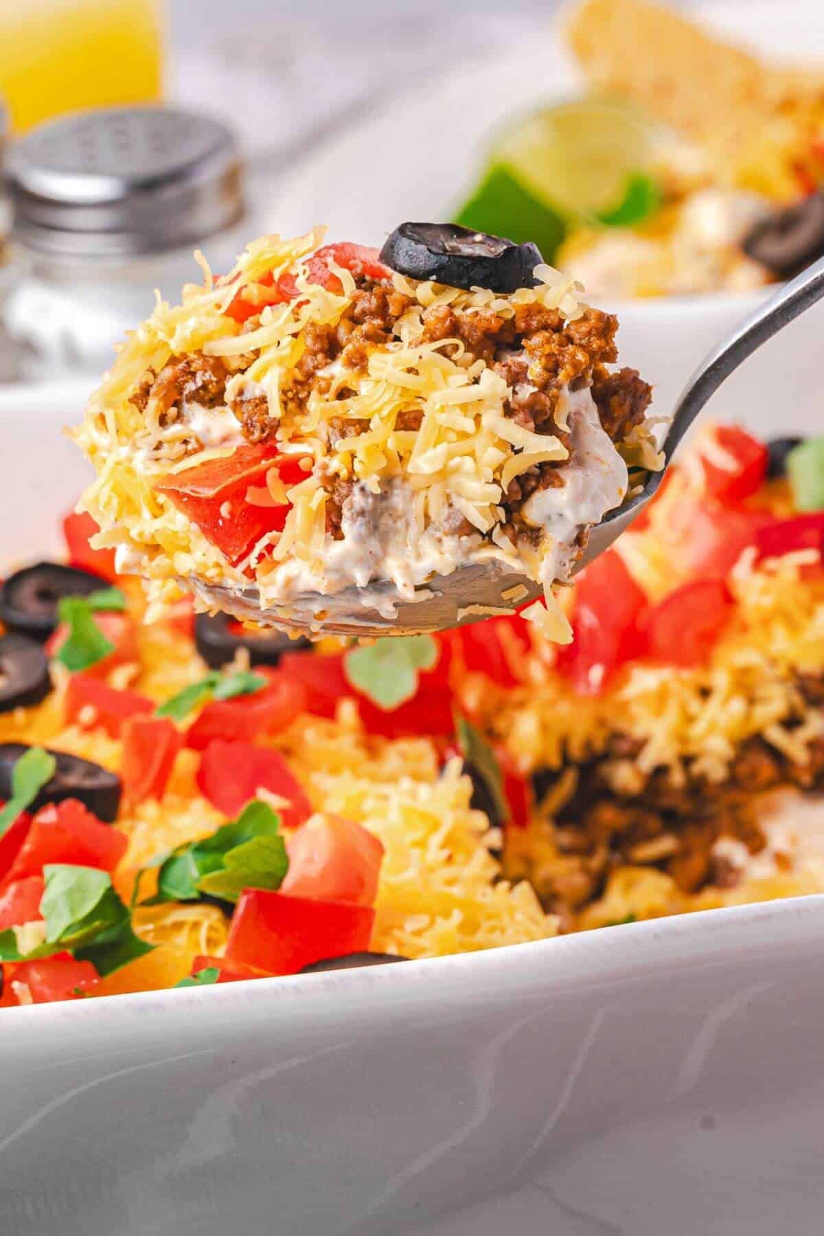A close-up of a spoonful of layered taco dip topped with shredded cheese, ground beef, chopped tomatoes, black olives, and fresh herbs.
