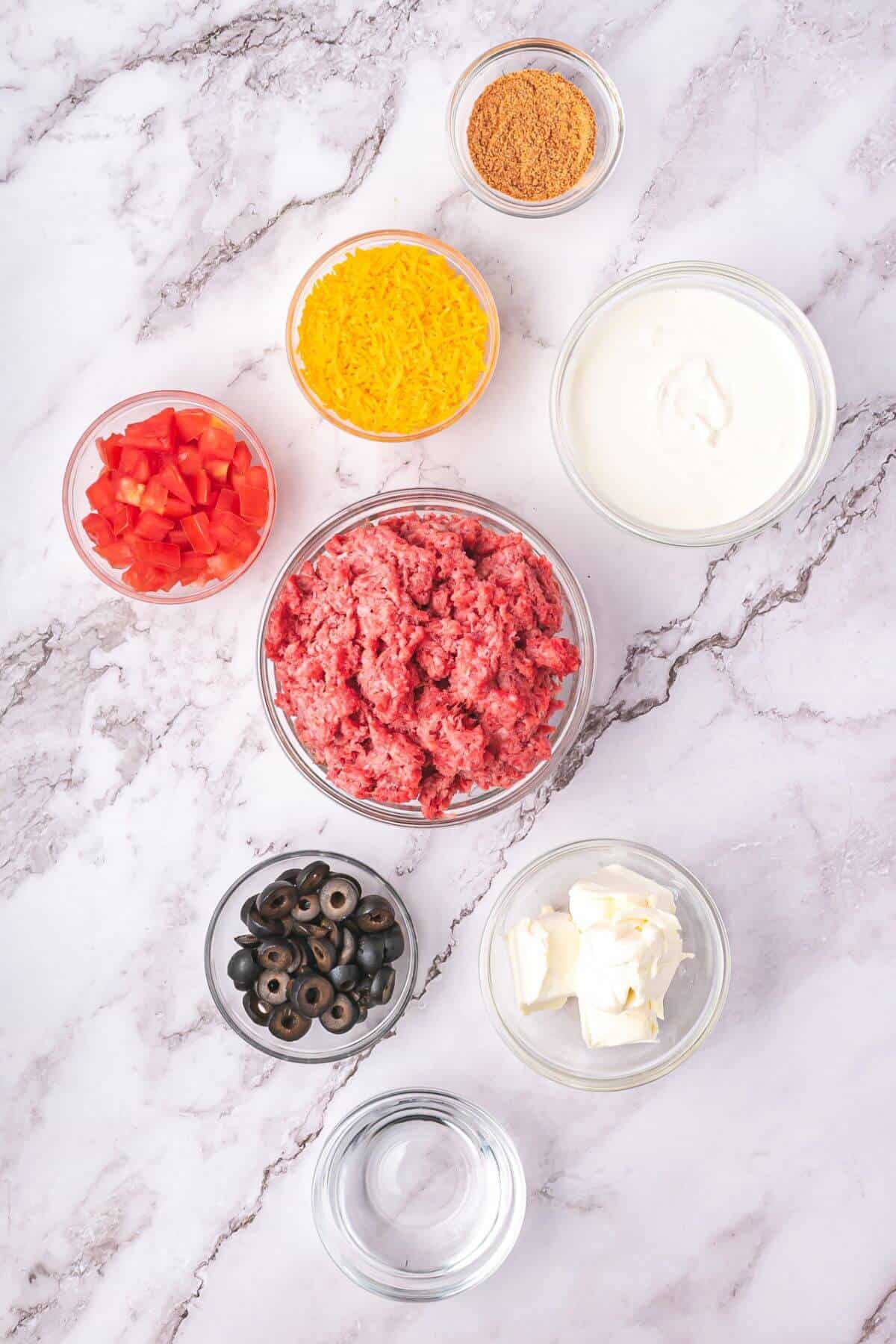 Bowls containing ground beef, shredded cheese, sour cream, diced tomatoes, taco seasoning, sliced black olives, cream cheese, and water arranged on a marble surface.