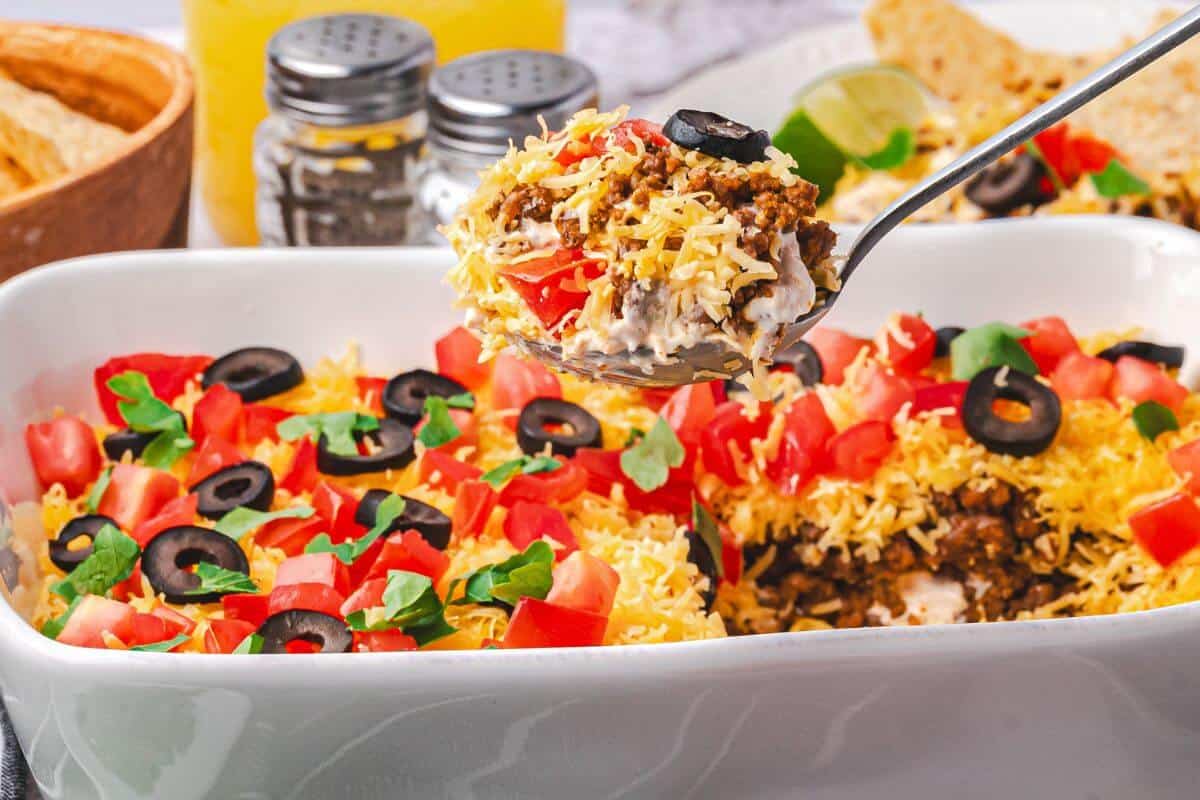A close-up of a layered taco dip in a white dish, topped with shredded cheese, diced tomatoes, olives, and greens, with a spoon lifting a portion.