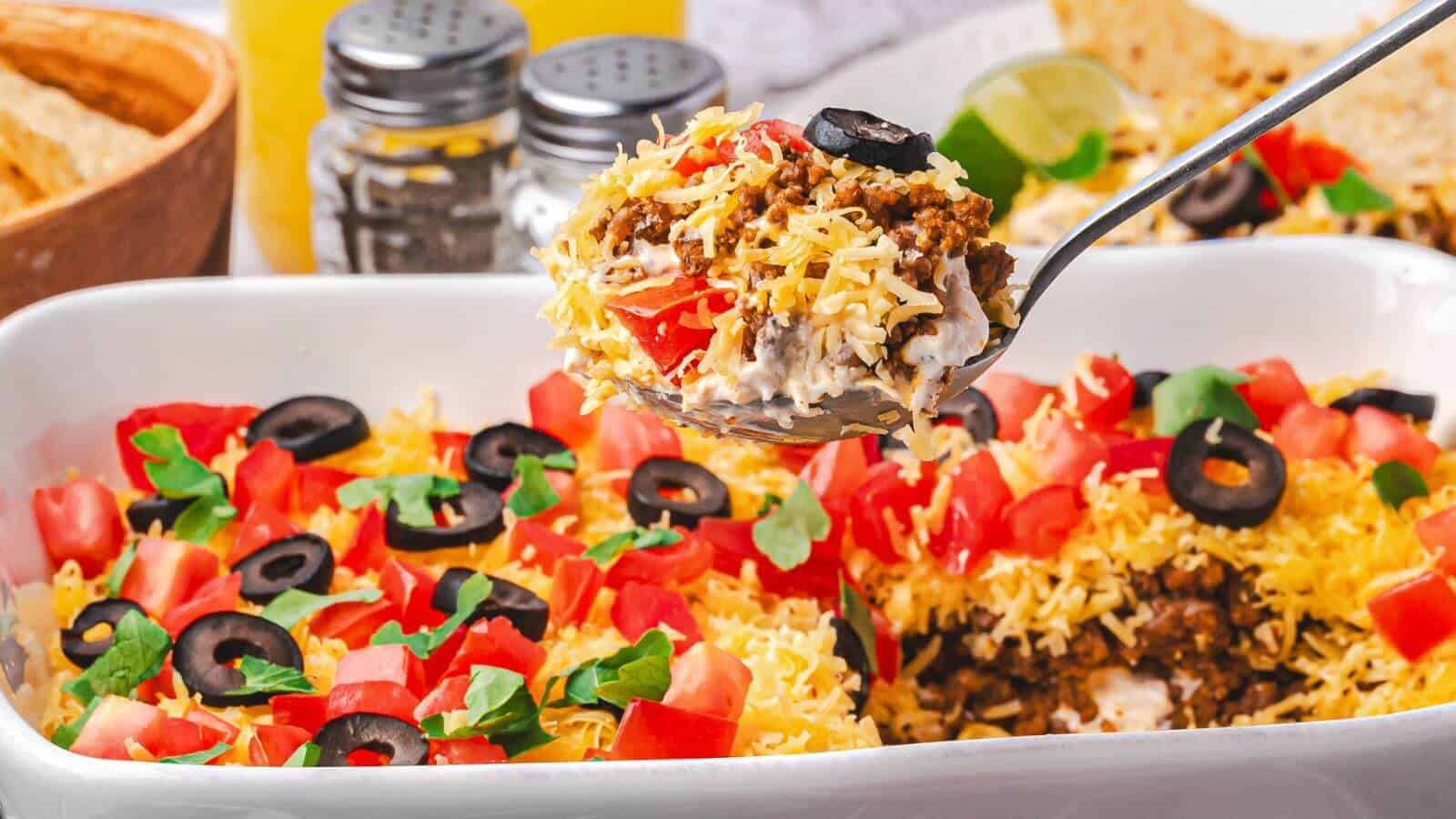A close-up of a layered taco casserole topped with shredded cheese, chopped tomatoes, black olives, and herbs, with a serving spoon lifting a portion.