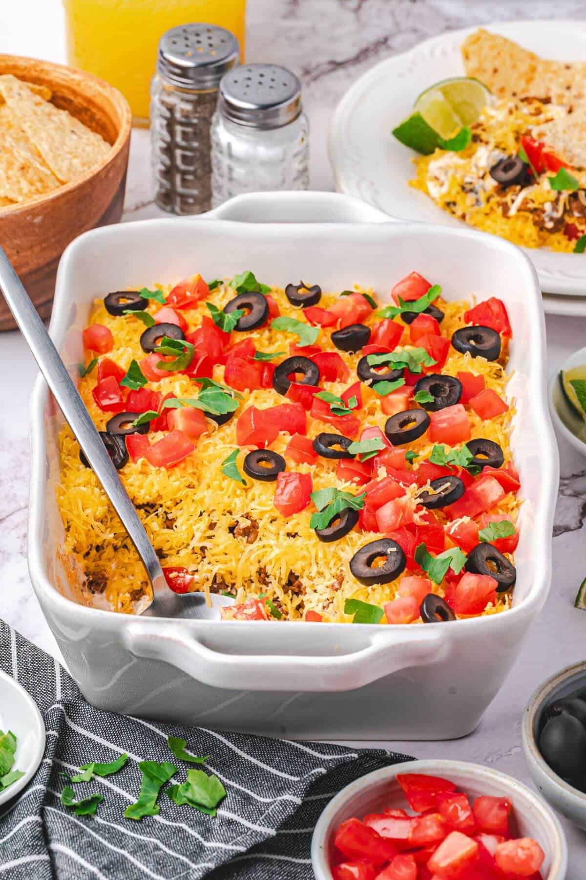 A white casserole dish filled with a layered dip topped with shredded cheese, chopped tomatoes, black olives, and lettuce, with a serving spoon inside. Tortilla chips and condiments are nearby.