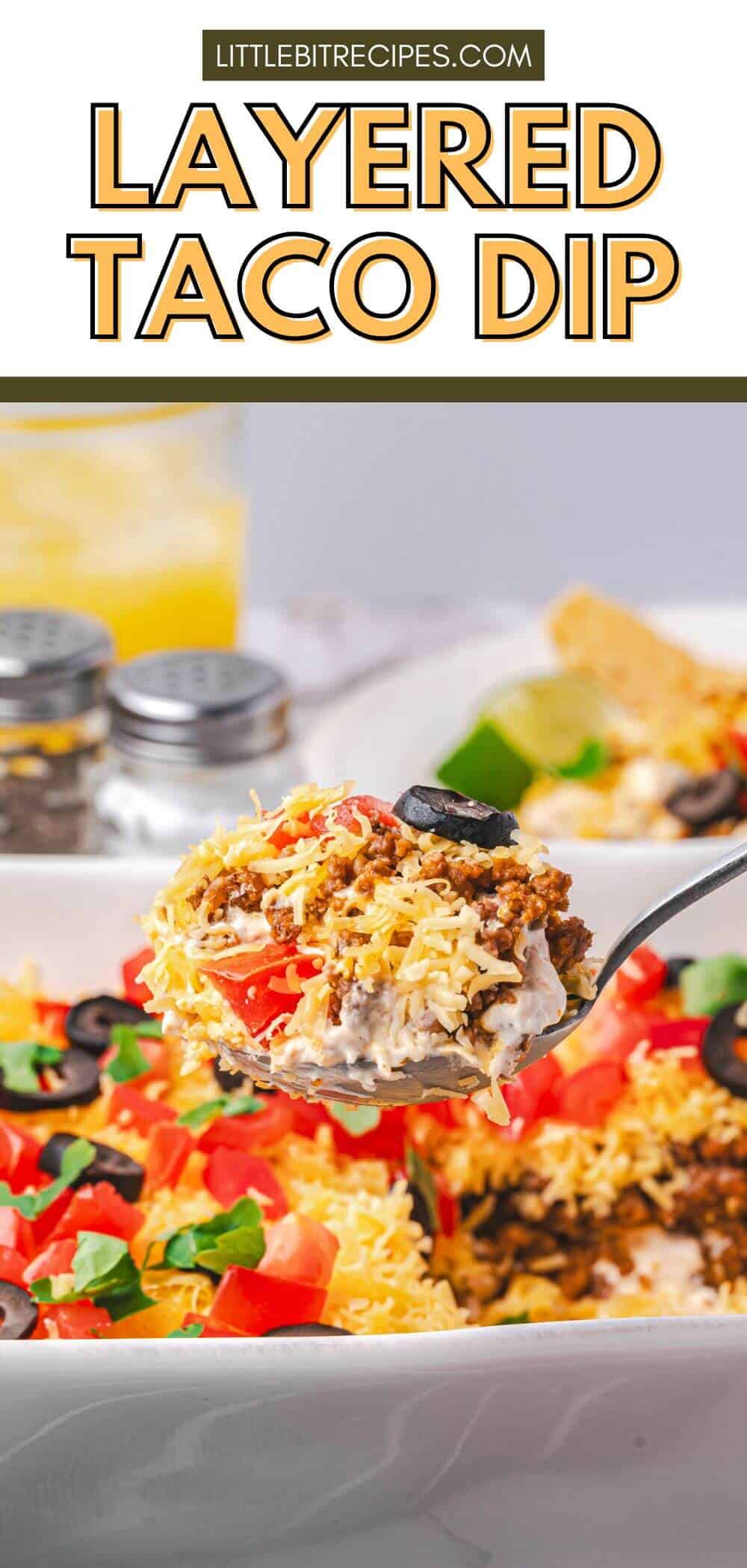 A spoonful of layered taco dip with ground meat, shredded cheese, tomatoes, olives, and lettuce held above a dish of the same dip.