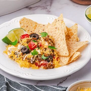 A plate with tortilla chips, ground beef dip topped with shredded cheese, tomatoes, olives, and fresh herbs, garnished with a lime wedge.