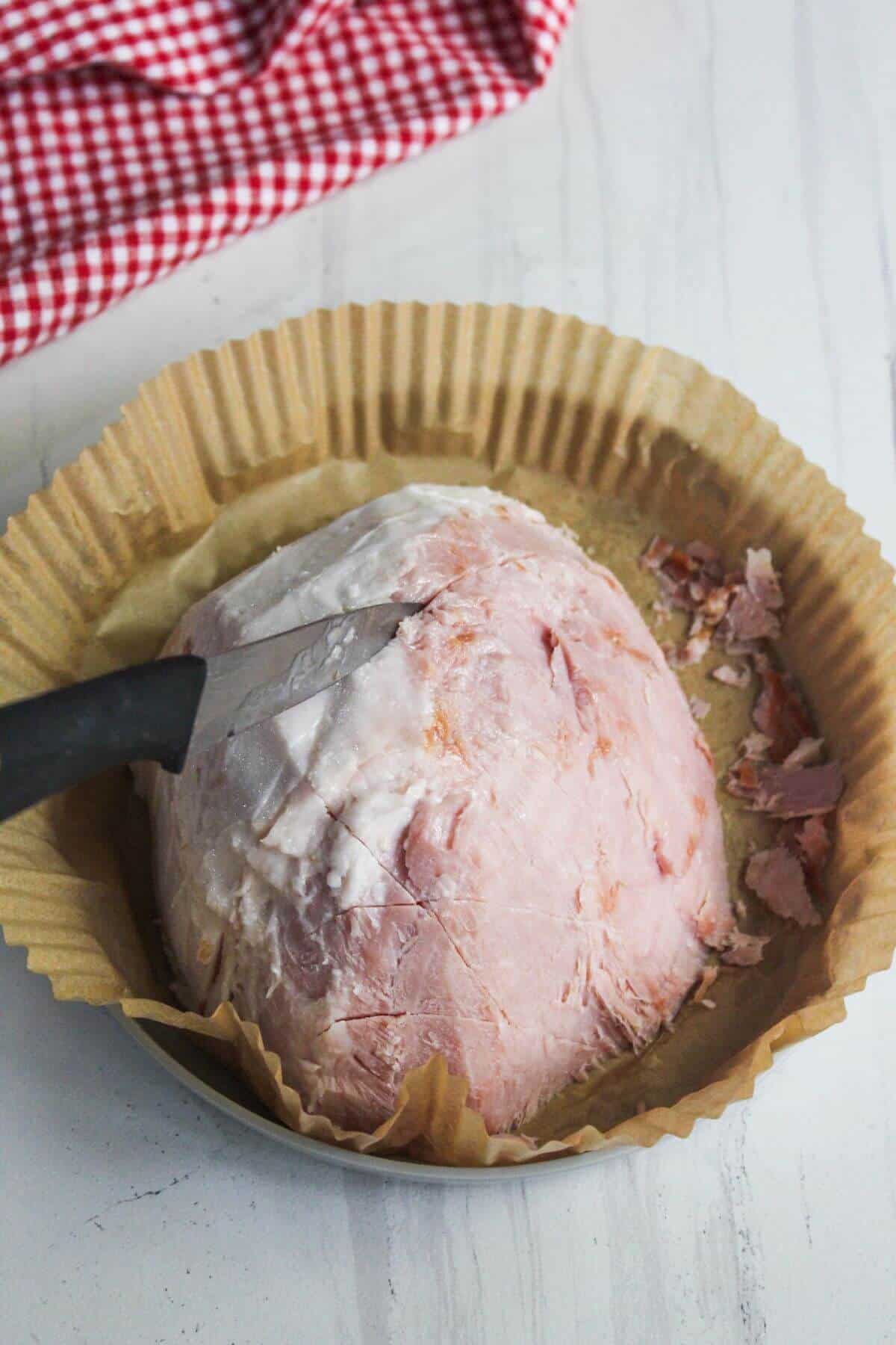 A knife inserted into a partially sliced ham placed in a round baking dish lined with parchment paper. A red and white checkered cloth is in the background.
