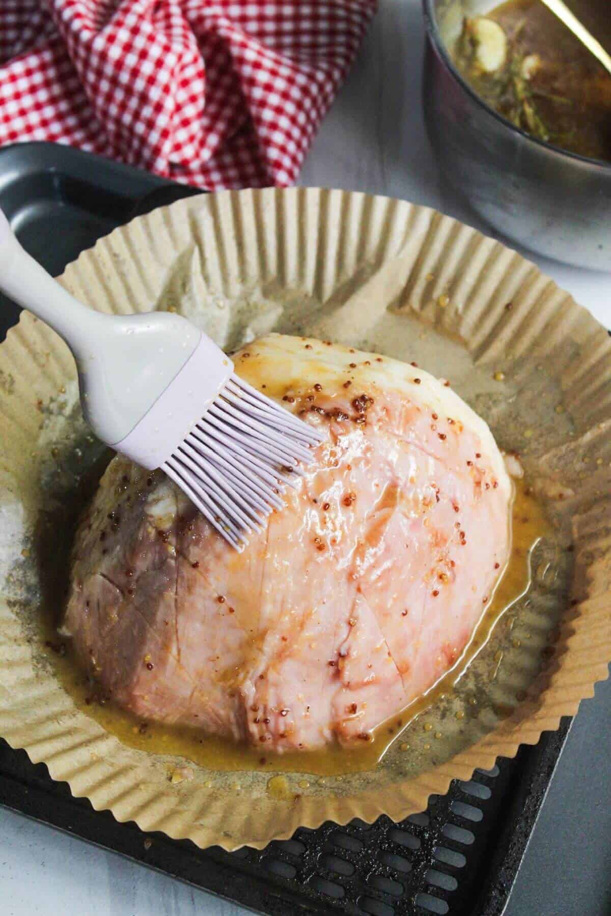 A brush applies marinade to a raw, seasoned ham placed on parchment paper in a baking dish. A bowl and a red-and-white checkered cloth are in the background.
