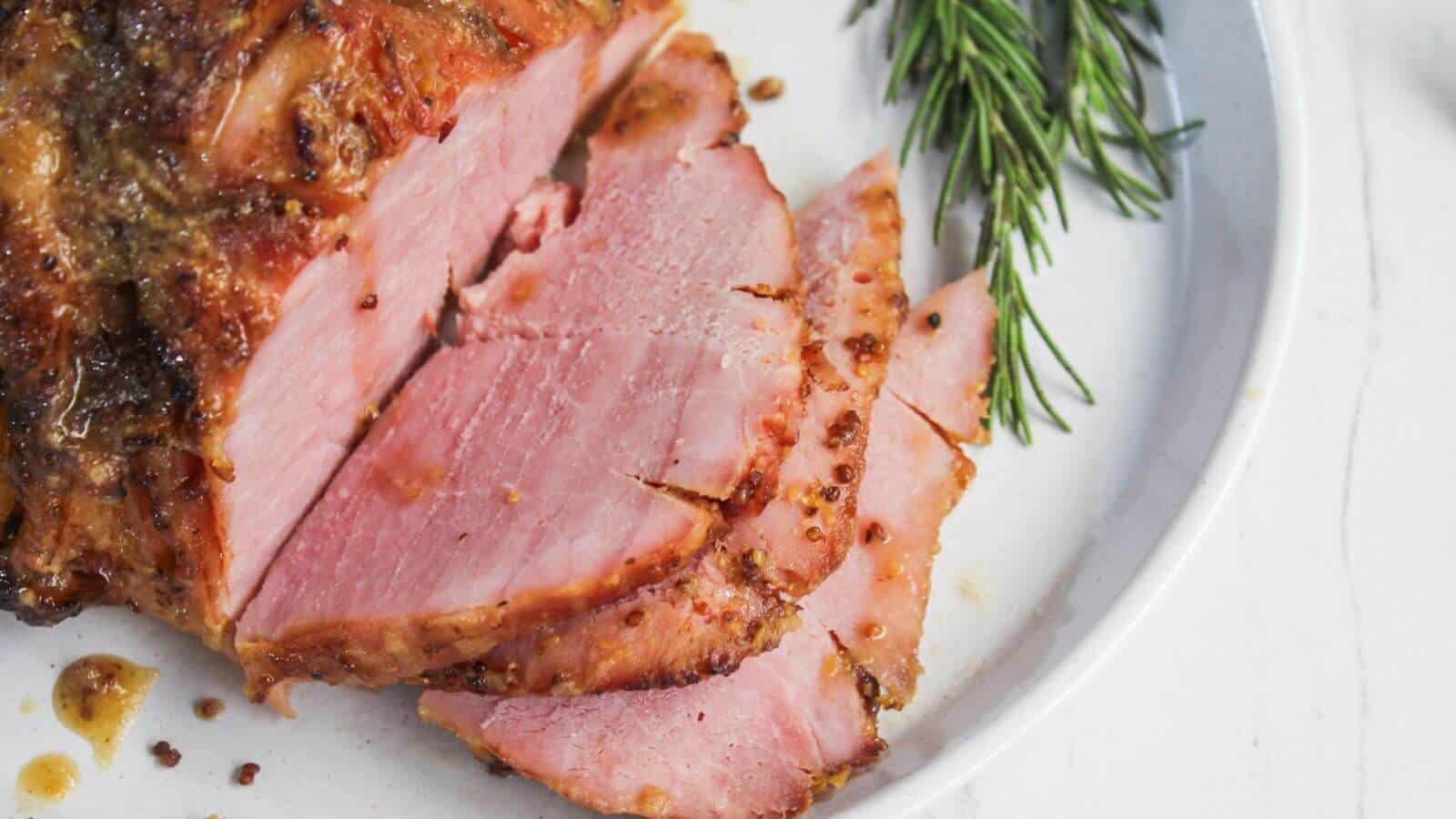 Sliced roasted ham garnished with sprigs of rosemary on a white plate.