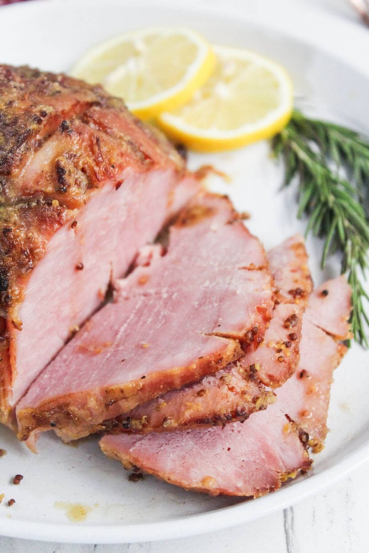 Sliced roasted ham on a plate garnished with lemon slices and rosemary sprigs.