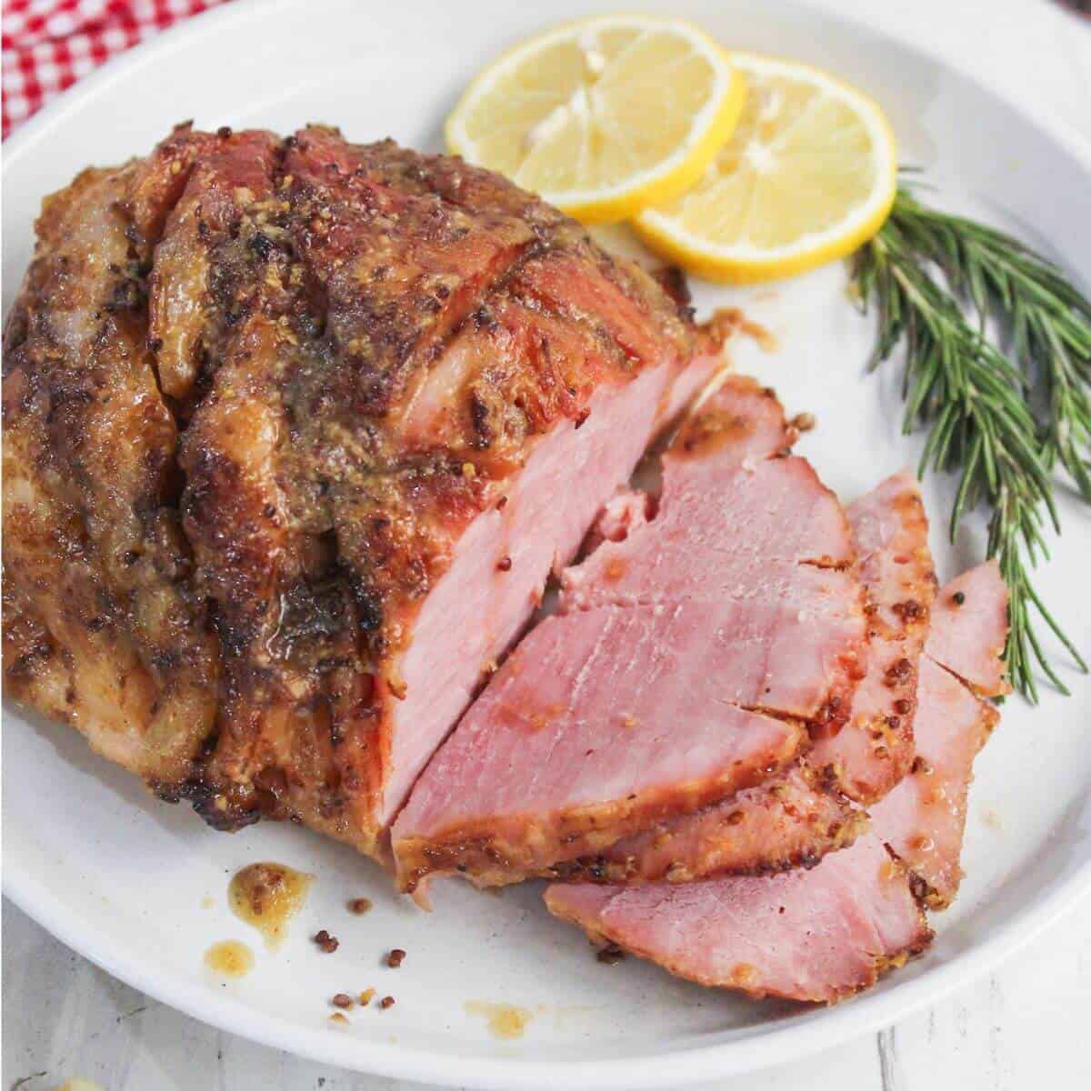 Sliced glazed ham on a plate garnished with two lemon slices and a sprig of rosemary.