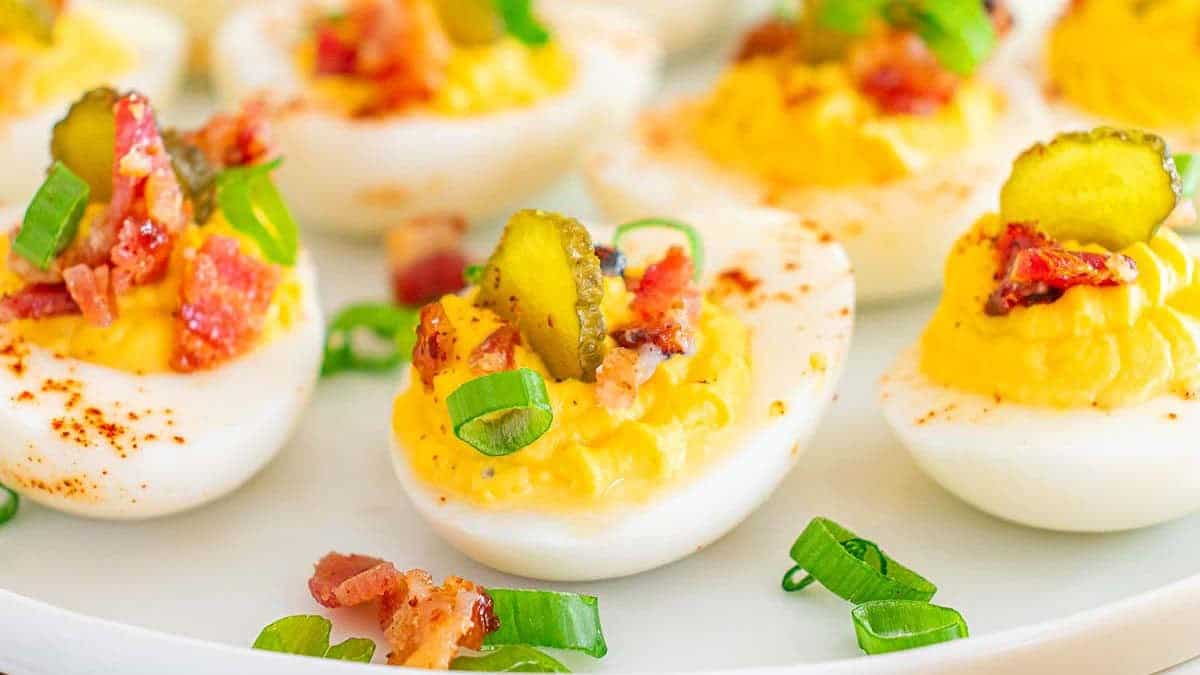 A plate full of deviled eggs with a sliced pickle and bacon on top.