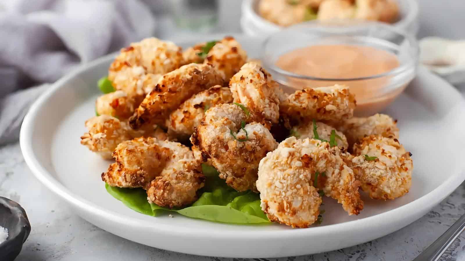 A white plate with breaded, baked shrimp on a bed of lettuce, served with a side of orange dipping sauce in a clear bowl.