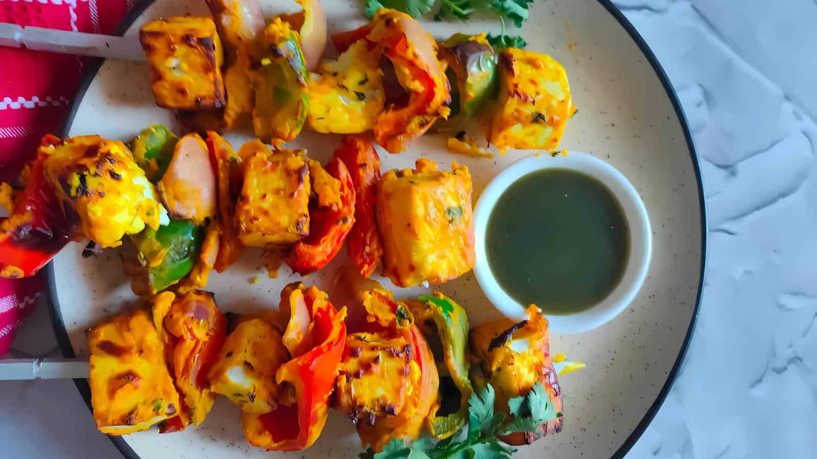 A plate with skewers of grilled paneer and vegetables, served with a small bowl of green dipping sauce and garnished with cilantro.