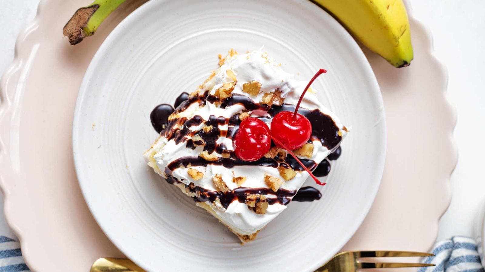 A slice of dessert topped with whipped cream, chocolate syrup, chopped nuts, and two cherries on a white plate, with a whole banana beside it.