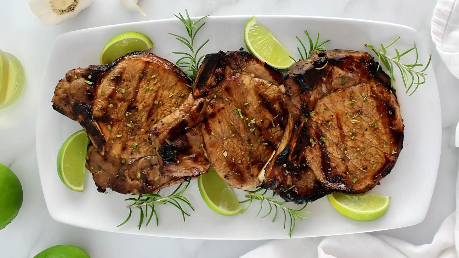 Three grilled pork chops garnished with rosemary and lime wedges are arranged on a white rectangular plate atop a marble surface.