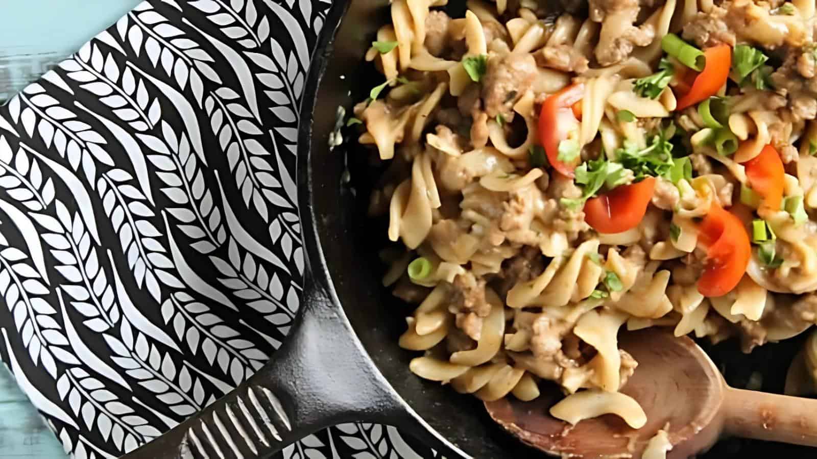 A cast iron skillet filled with pasta, ground meat, chopped tomatoes, and herbs sits on a patterned black and white cloth with a wooden spoon resting inside.