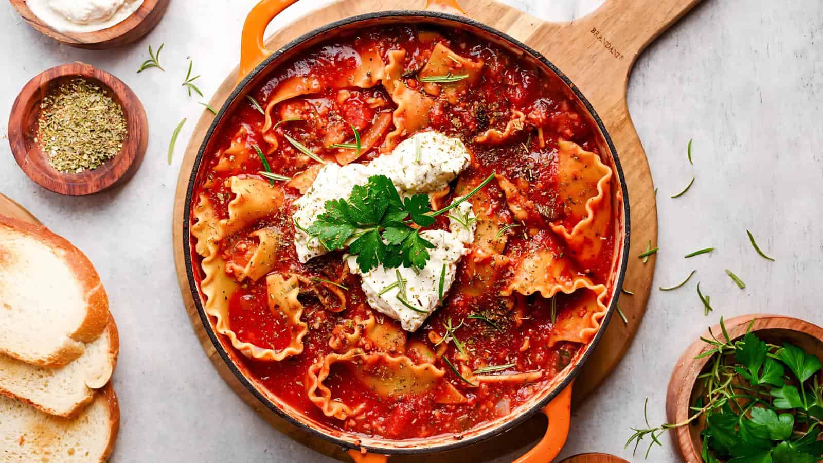 A skillet filled with lasagna noodles in tomato sauce, topped with dollops of ricotta cheese and fresh parsley, surrounded by slices of bread and small bowls of herbs.