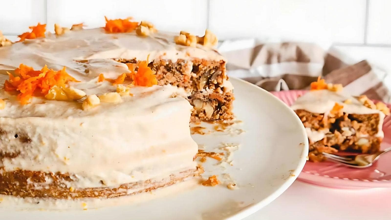 A round carrot cake with white frosting, topped with chopped nuts and grated carrot, sits on a plate with a slice served on a pink dish in the background.