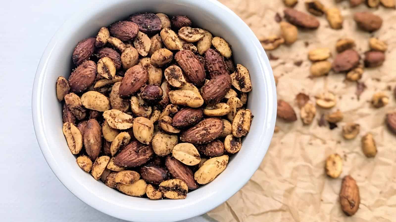 A white bowl filled with seasoned mixed nuts, with more nuts scattered on a piece of brown parchment paper beside it.