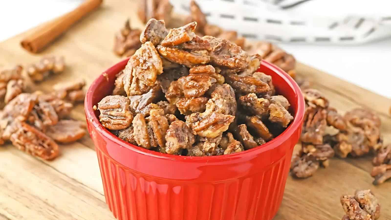 A red ramekin filled with candied pecans sits on a wooden surface, with additional pecans scattered around it.