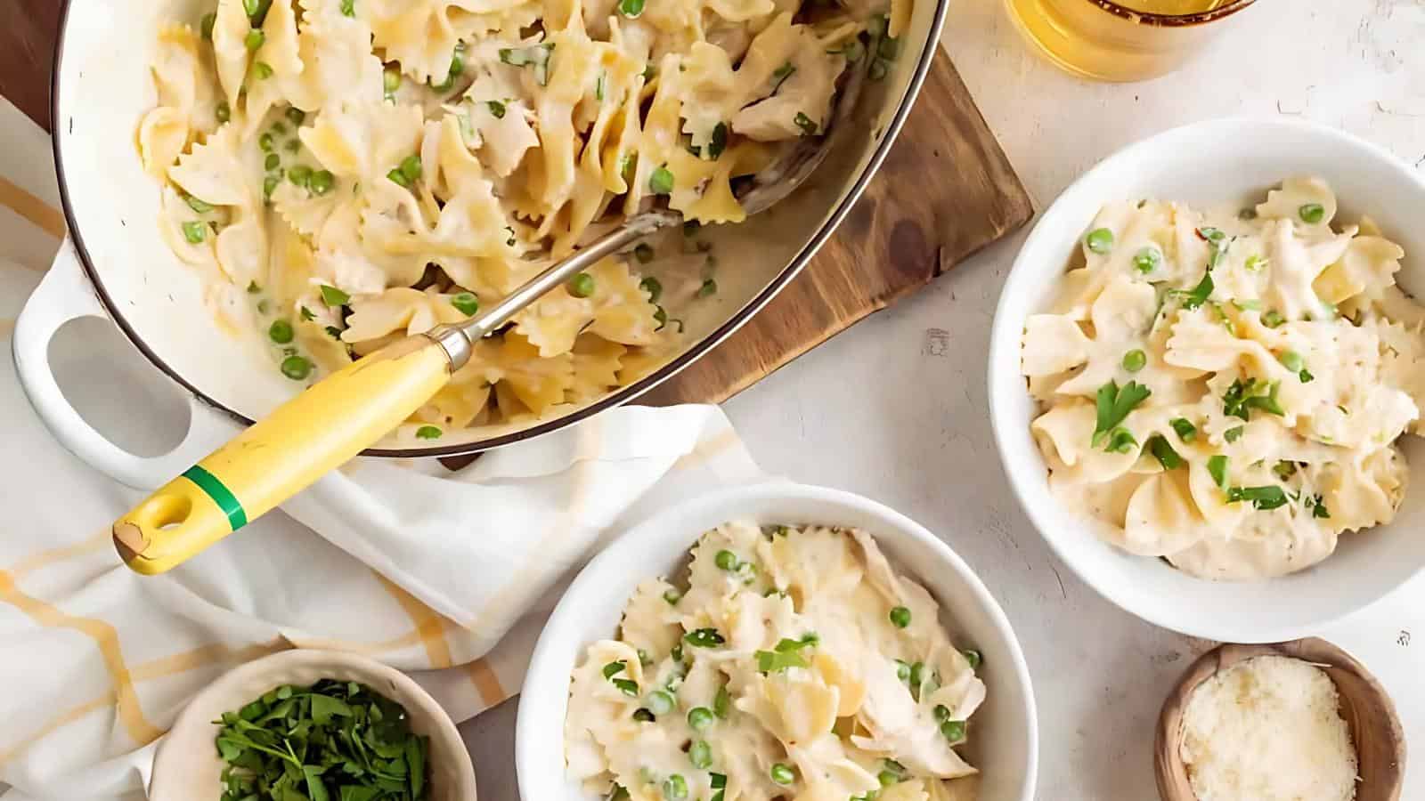 A pot and two bowls of bow-tie pasta with creamy sauce and peas, garnished with herbs. A small bowl of chopped herbs and grated cheese are on the side.