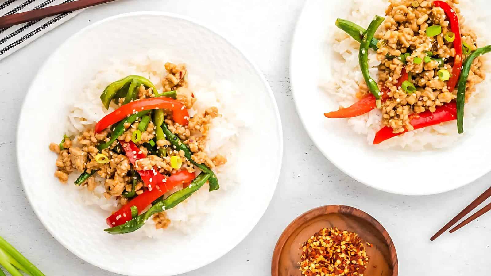 Two plates of white rice topped with stir-fried ground meat, red bell peppers, green beans, and garnished with sesame seeds and chopped green onions, with a dish of crushed red pepper on the side.