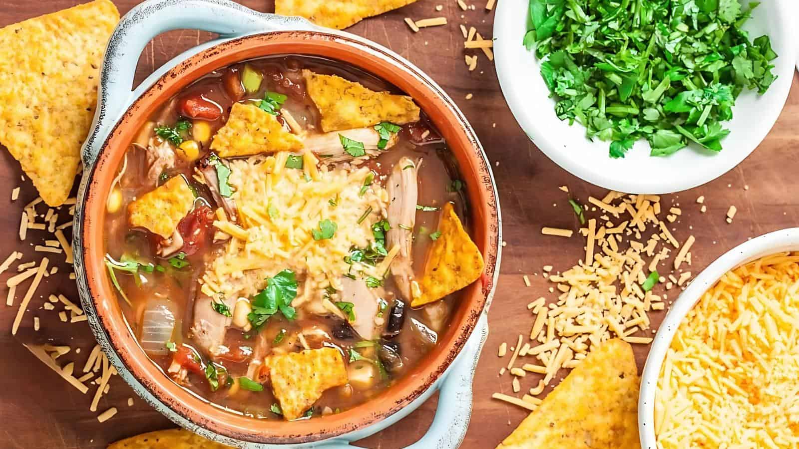 A bowl of chili topped with shredded cheese, tortilla chips, and chopped cilantro, surrounded by extra chips, shredded cheese, and a bowl of cilantro on a wooden surface.