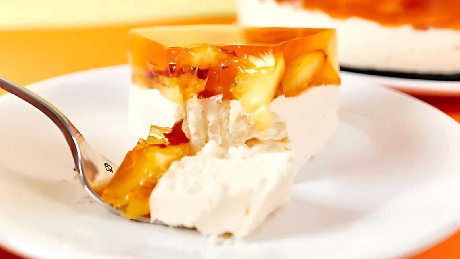 A slice of layered dessert with white cream and orange jelly topping on a white plate, with a fork taking a bite.