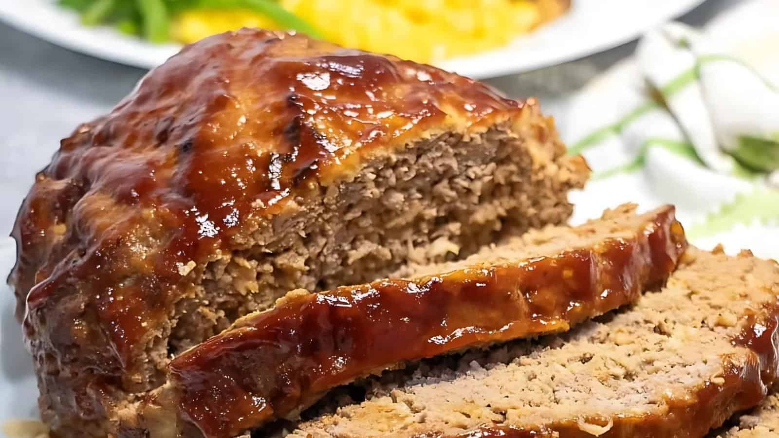 A glazed meatloaf is sliced to show its interior, with a glossy brown coating and tender, moist texture. A plate with yellow and green food is visible in the background.