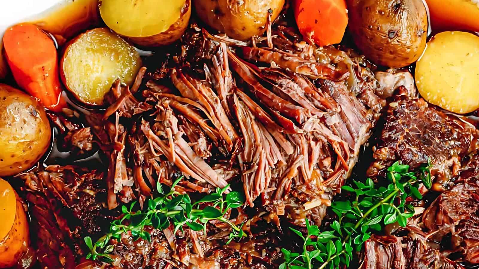 Shredded pot roast surrounded by whole potatoes, carrots, and fresh herbs, arranged on a serving dish.