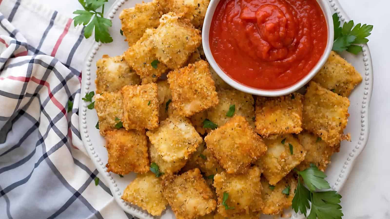 A plate of breaded, fried ravioli is served with a bowl of marinara sauce and garnished with parsley. A plaid napkin is on the side.