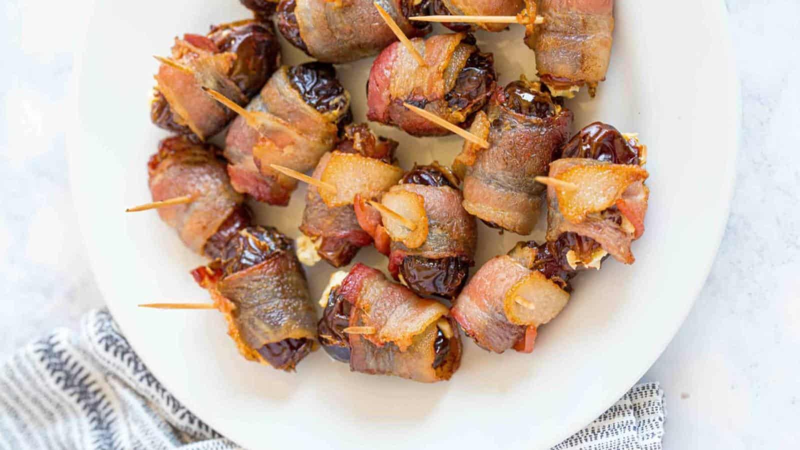 A white plate with several bacon-wrapped dates secured with toothpicks, arranged in a circular pattern.
