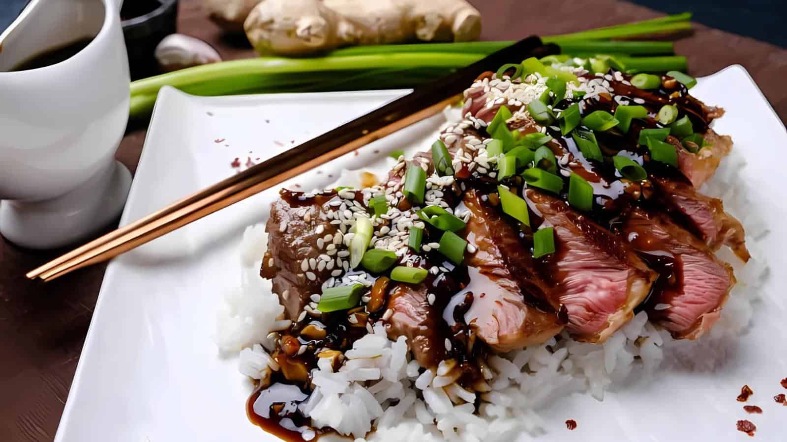 Sliced steak with teriyaki sauce, sesame seeds, and chopped green onions served over white rice on a square plate with chopsticks and a sauce dish.