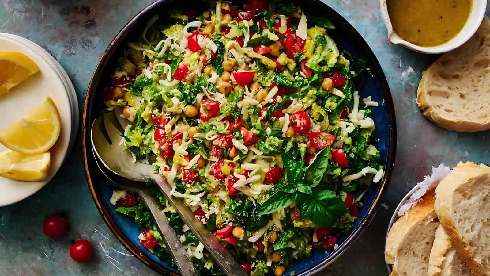 A bowl of chopped salad with lettuce, cherry tomatoes, cheese, chickpeas, and herbs, served with lemon wedges, bread slices, and a small jug of dressing.