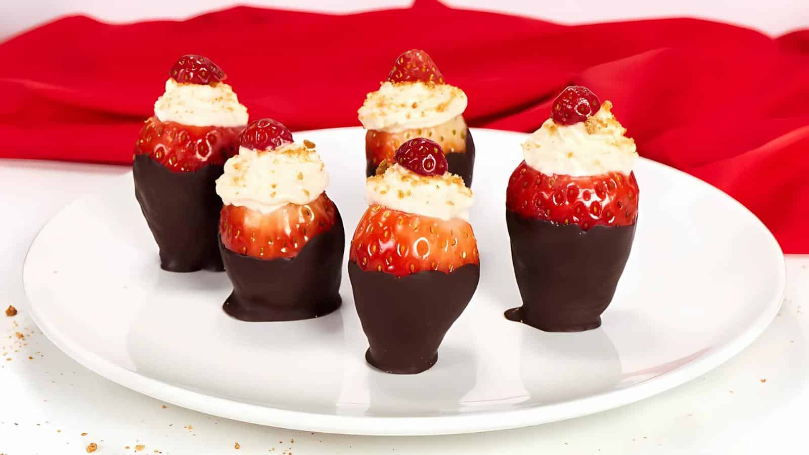 Five strawberries dipped in chocolate, topped with cream and a small berry, arranged on a white plate with a red cloth in the background.