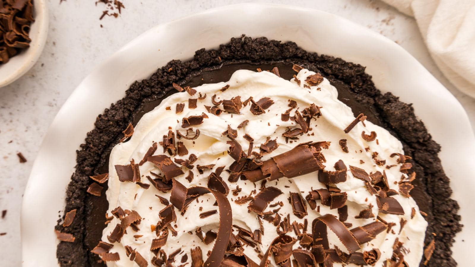 A chocolate cream pie with a cookie crust, topped with whipped cream and chocolate shavings, shown in a white pie dish from an overhead view.