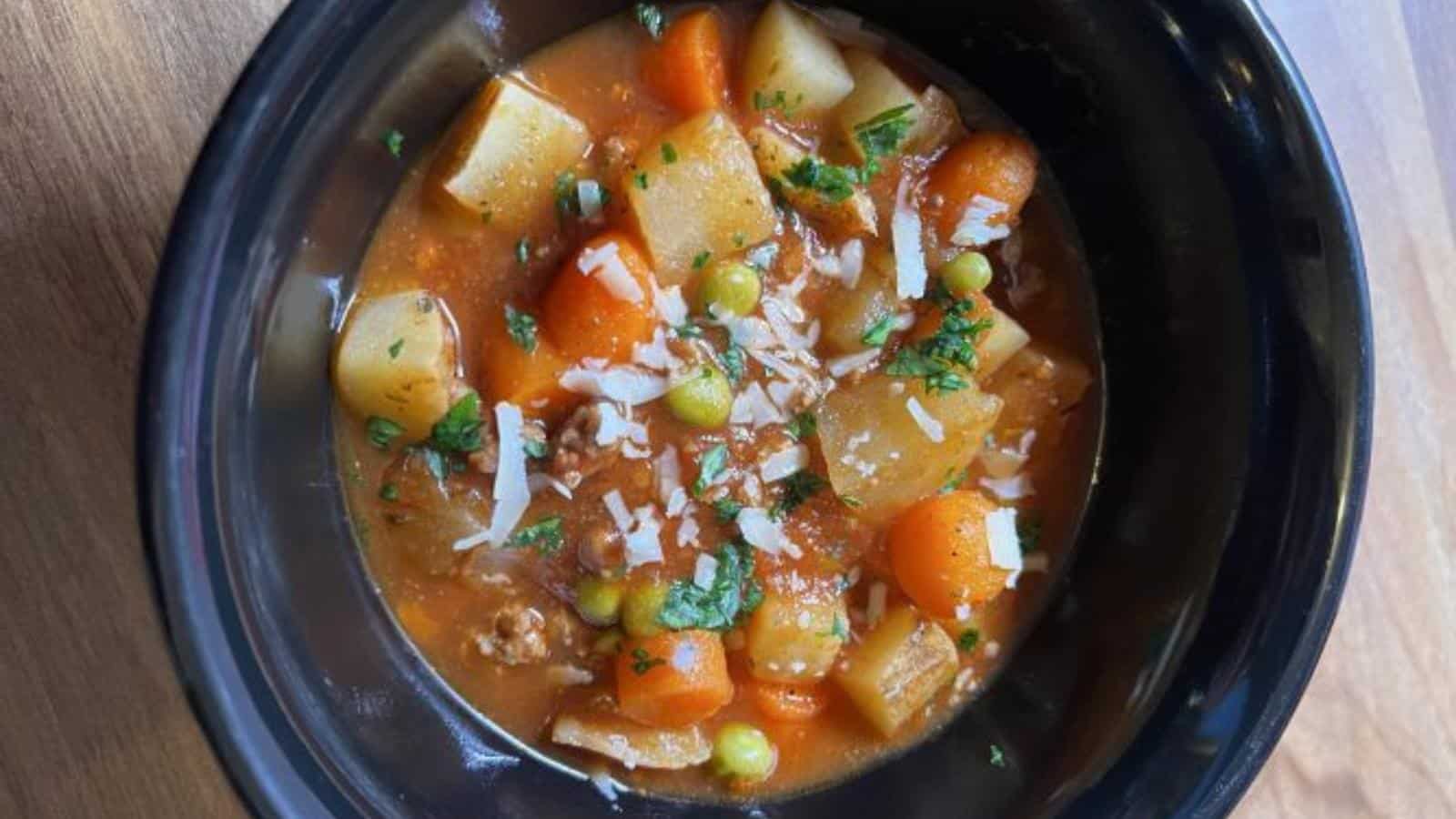A black bowl filled with vegetable stew containing carrots, potatoes, peas, ground meat, tomato-based broth, and garnished with chopped herbs and shredded cheese.