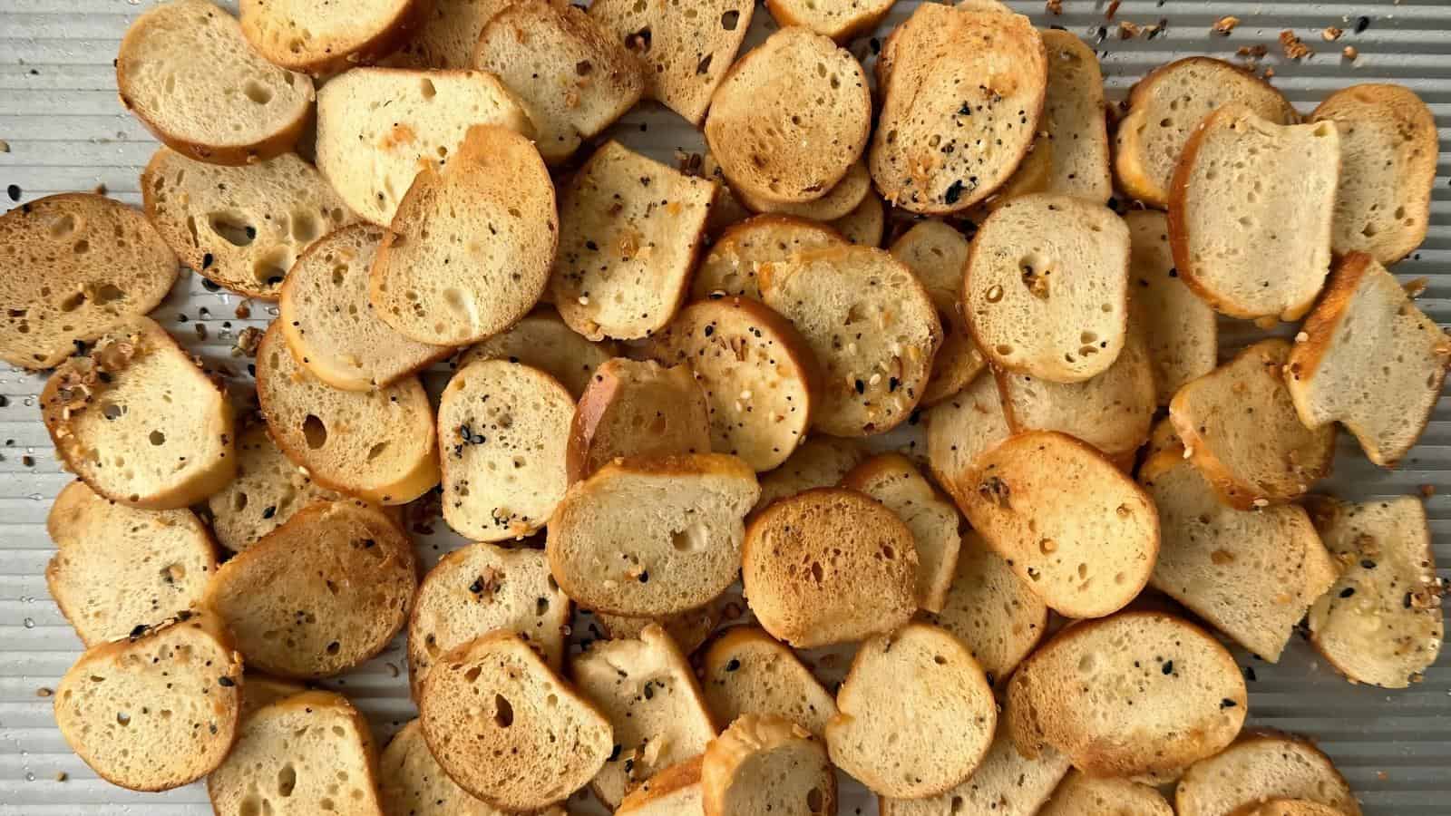 Scattered slices of toasted baguette chips with various seasonings on a ridged baking sheet.