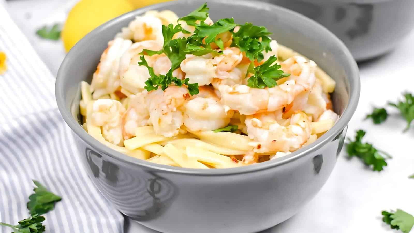 A gray bowl filled with pasta topped with shrimp and garnished with fresh parsley, with a striped cloth and lemon in the background.
