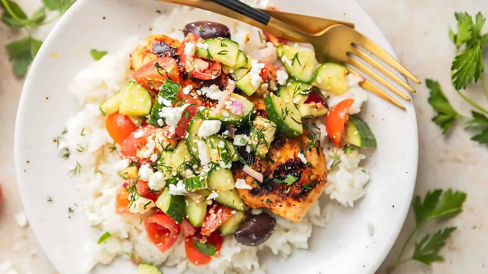 A plate of white rice topped with grilled chicken, chopped cucumber, tomato, olives, feta cheese, and herbs, with a fork and knife on the side.