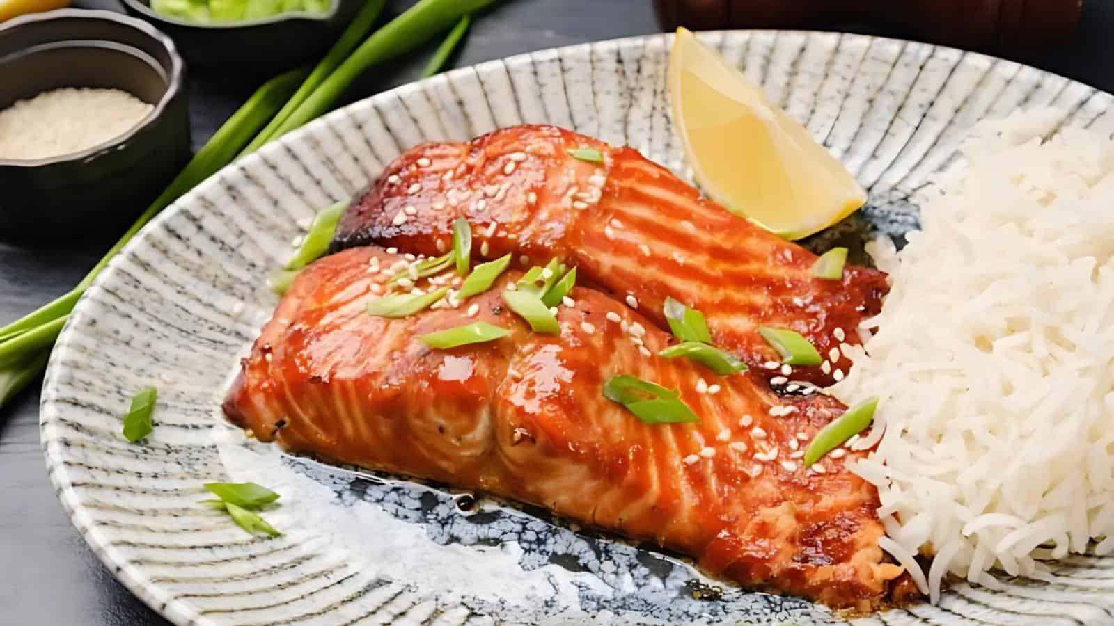 Grilled salmon fillets topped with sesame seeds and chopped green onions, served with a wedge of lemon and a side of white rice on a ceramic plate.